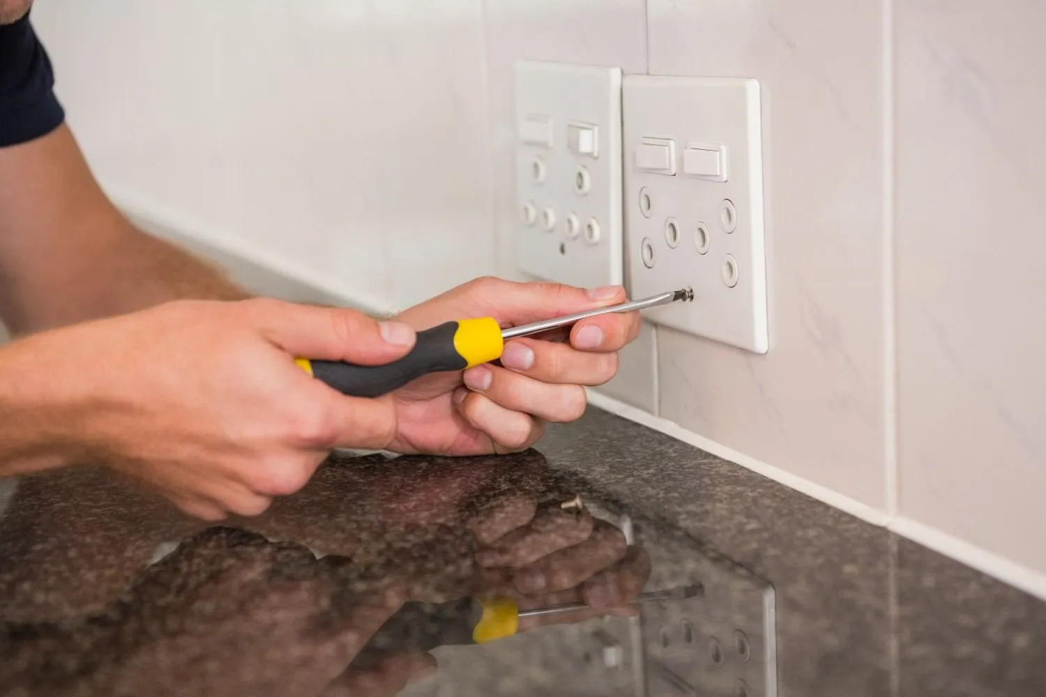Hands using a screwdriver on a wall outlet above a kitchen counter